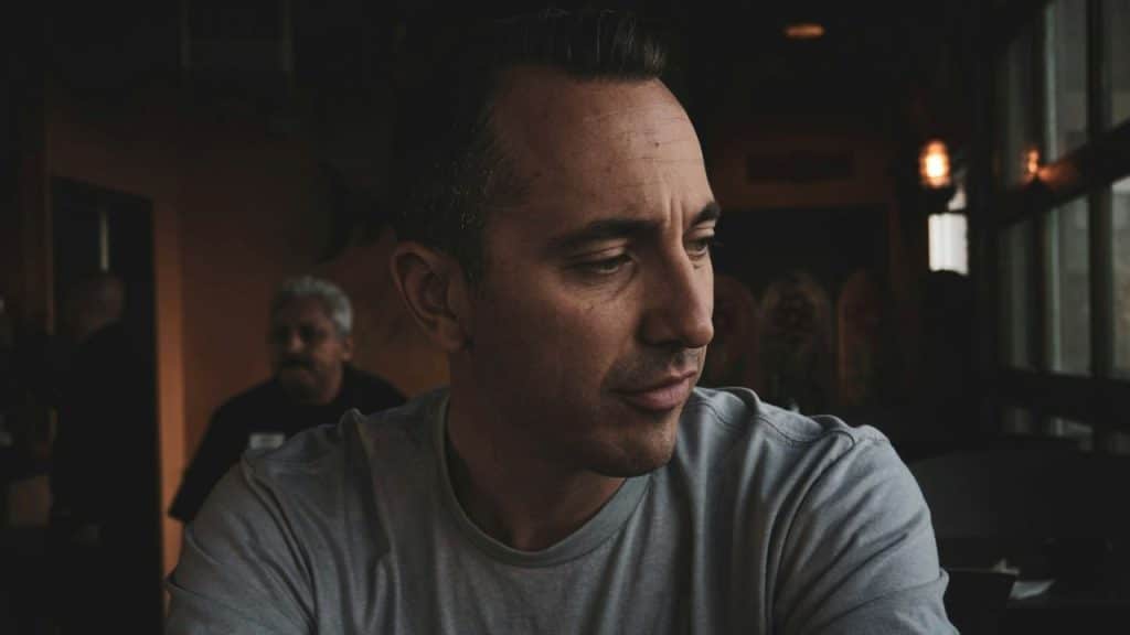A man in a grey t-shirt looks downward while sitting in a dimly lit indoor setting.