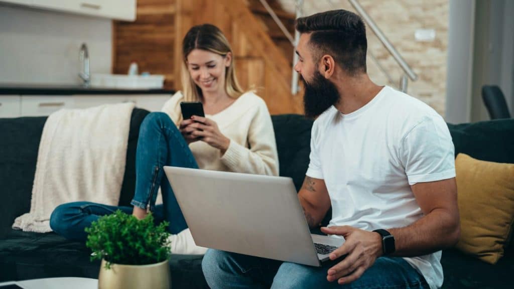 A man with a laptop and a woman with a phone sit on a sofa.