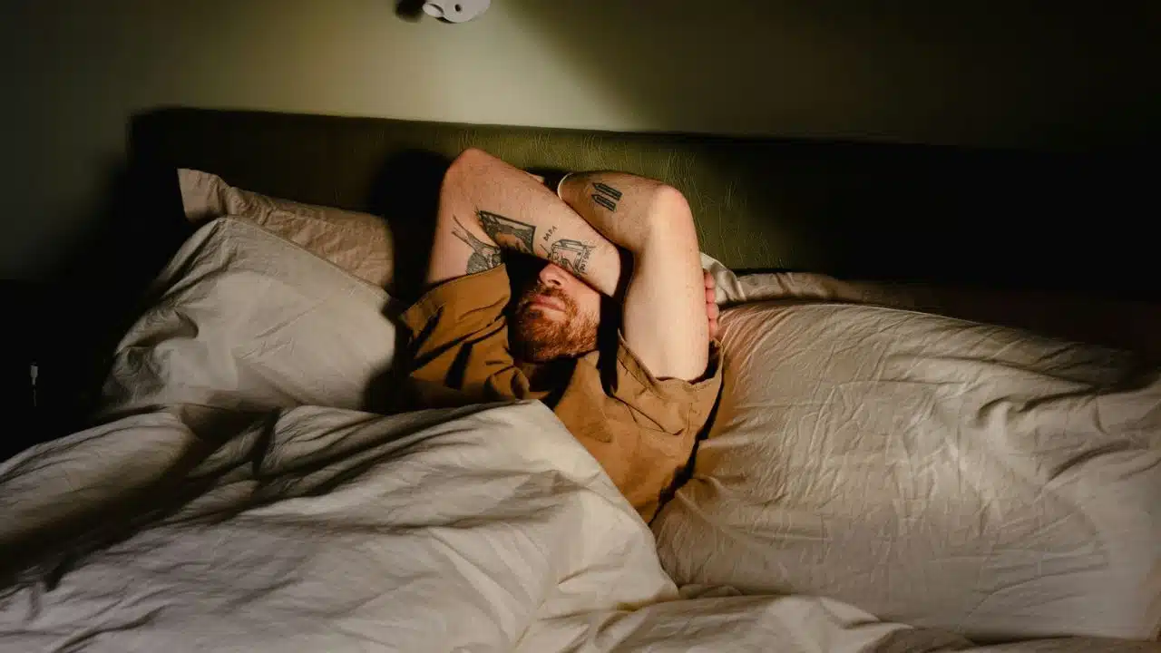 A man with tattoos on his arms lies in bed, covering his eyes with his forearm.