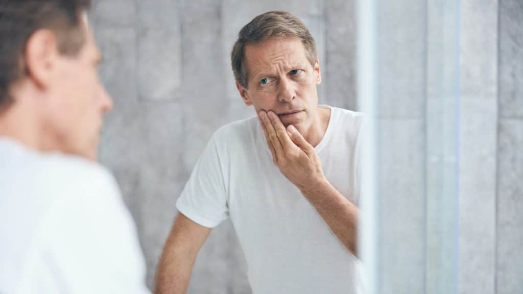 A middle-aged man in a white t-shirt looks at his reflection in a bathroom mirror.