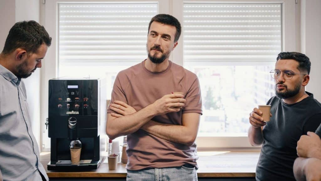 Three men stand in a kitchen holding small paper cups and looking in different directions.
