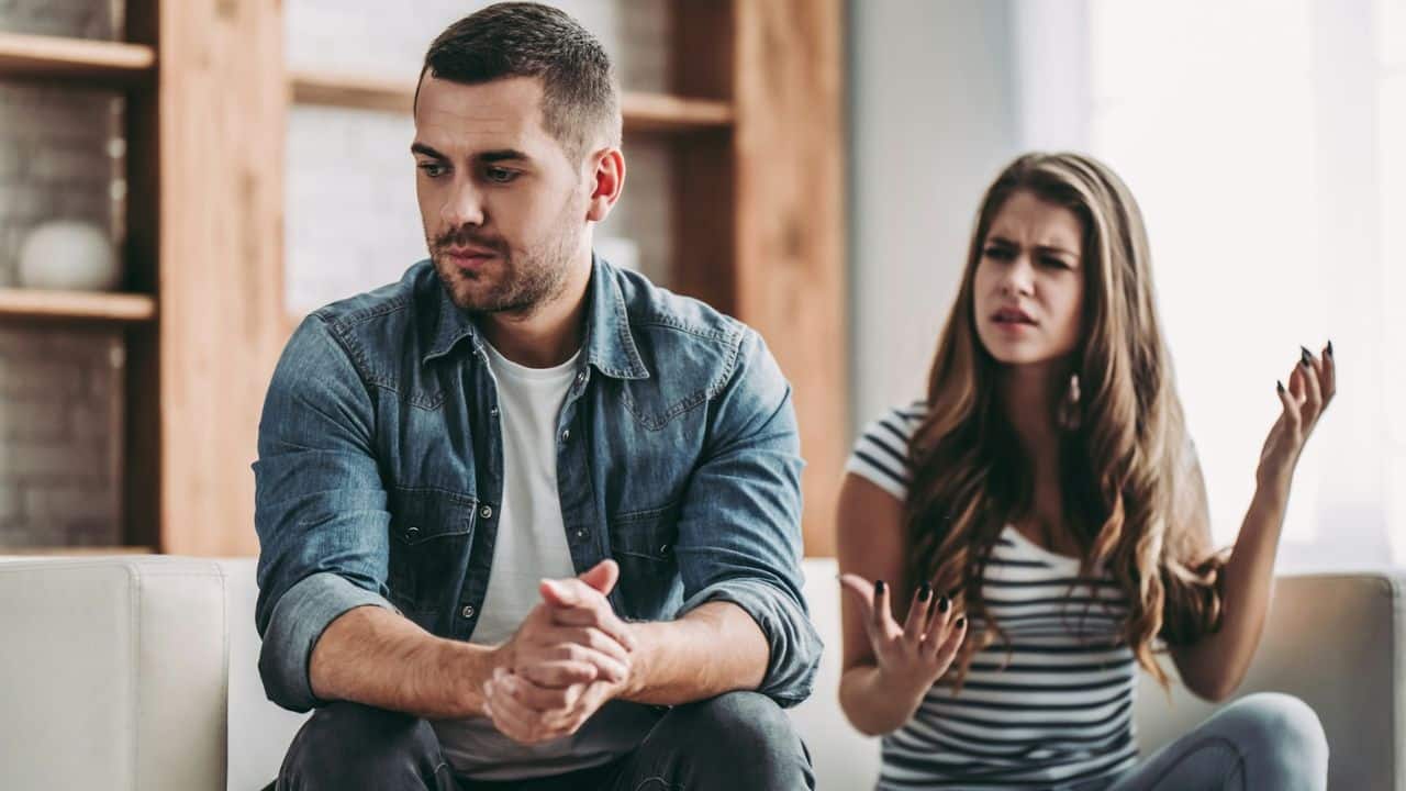 A man sits looking down while a woman behind him gestures with frustrated facial expressions.