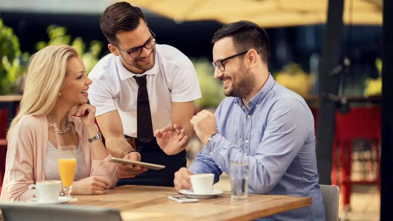 A couple smiling as a waiter shows them a menu at an outdoor café.