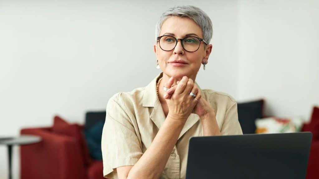 A woman with short grey hair and glasses looks thoughtfully away with hands clasped.