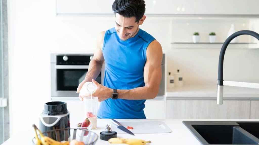 A man in a blue tank top pouring liquid into a blender cup with fruit.