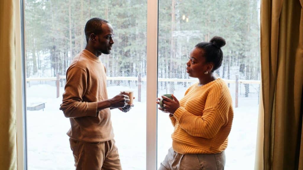 Two people holding mugs stand talking indoors by a large window overlooking a snowy forest.
