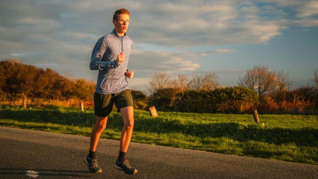 A young man in athletic wear runs on a paved road during a golden sunset.