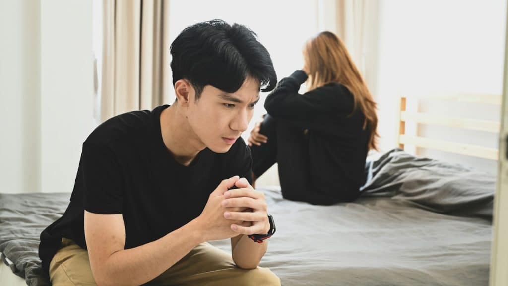 An upset man is sitting on the corner of a bed while a woman sits on the bed in the background.