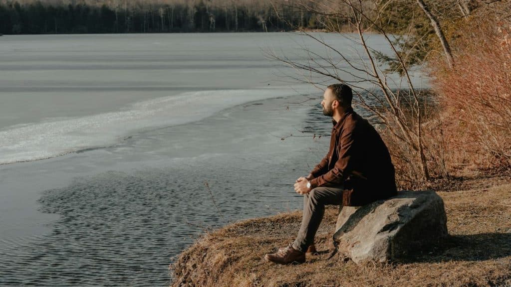 A bearded man sits on a rock overlooking a calm, partially frozen lake in winter.
