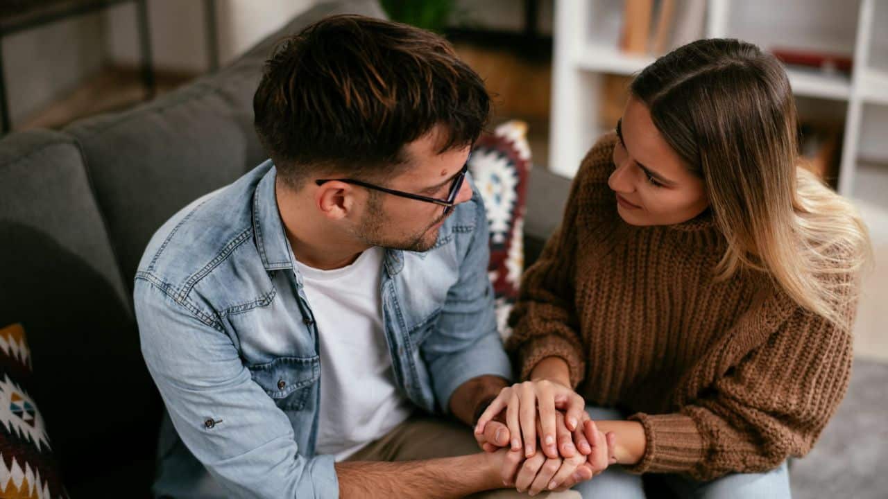 A man and woman sit close together on a sofa holding each other's hands.