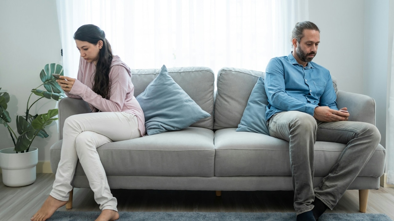 A man and woman sit on opposite ends of a sofa, both looking at smartphones.