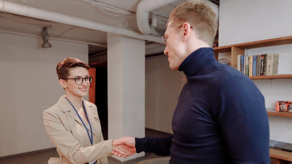 A woman in a beige coat and glasses smiling while shaking hands with a man in a navy blue turtleneck in an office or library setting.