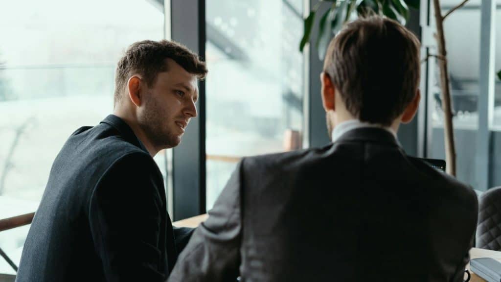 Two men in suits sit at a table in a modern office, engaged in a conversation.