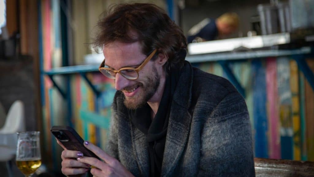A man with glasses and a beard smiles while looking down at a mobile phone.