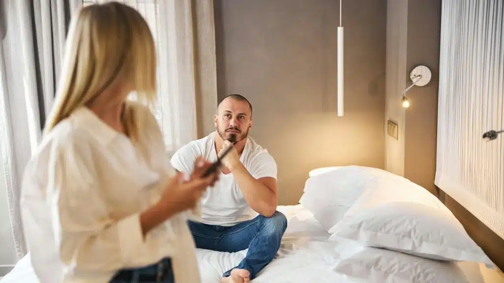 A man sits on a bed looking thoughtfully at a woman holding a phone.