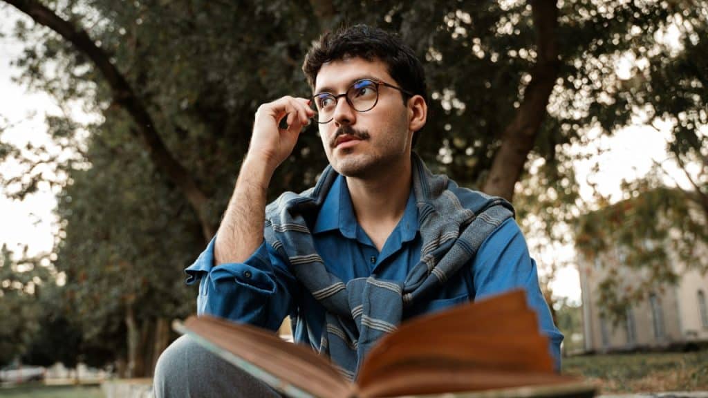 A man with a mustache and glasses sits outside with an open book, looking upward.