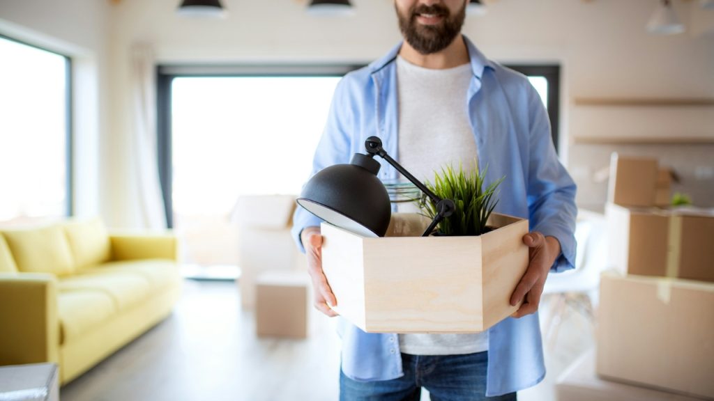 A man holds a wooden crate containing a lamp and plant in a bright room.