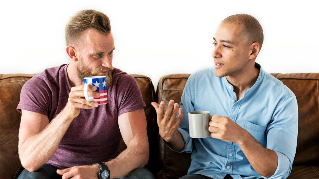 Two men sit on a leather sofa and talk while holding coffee mugs.