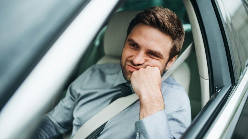 A man in a dress shirt sits in a car leaning his face on his hand.