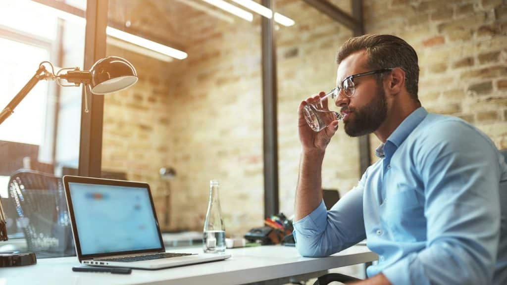 A bearded man in a blue shirt drinks water at a desk with a laptop.