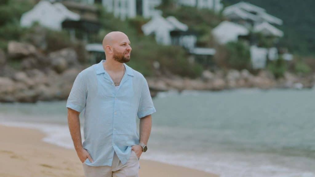 A bald man with a beard walks on a beach wearing a light blue button-up shirt.