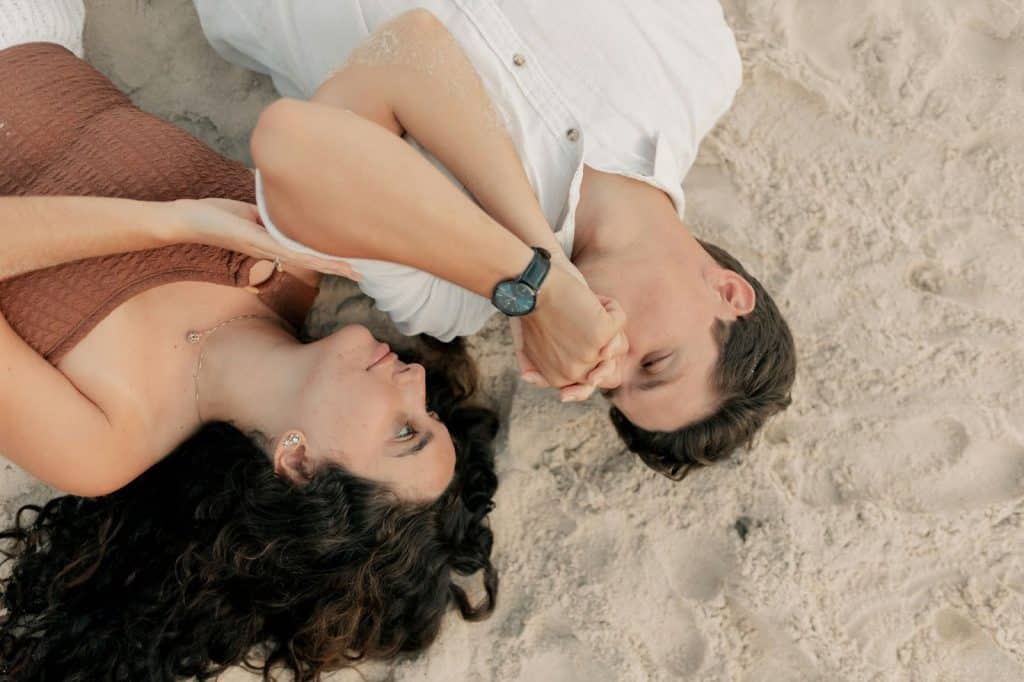 A man and woman lying on the sand while the man kisses the woman’s hand.