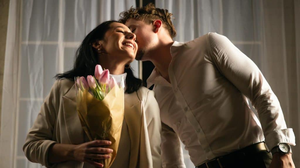 A man whispers to a smiling woman holding a bouquet of pink tulips.