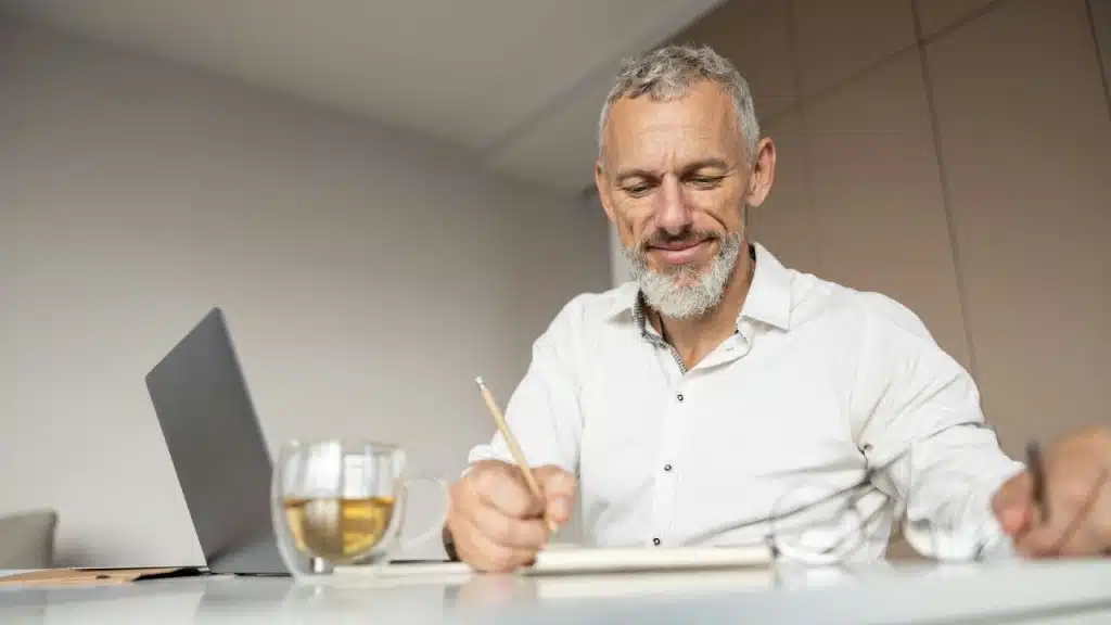 A smiling, gray-bearded man in a white shirt writes in a notebook near a laptop.