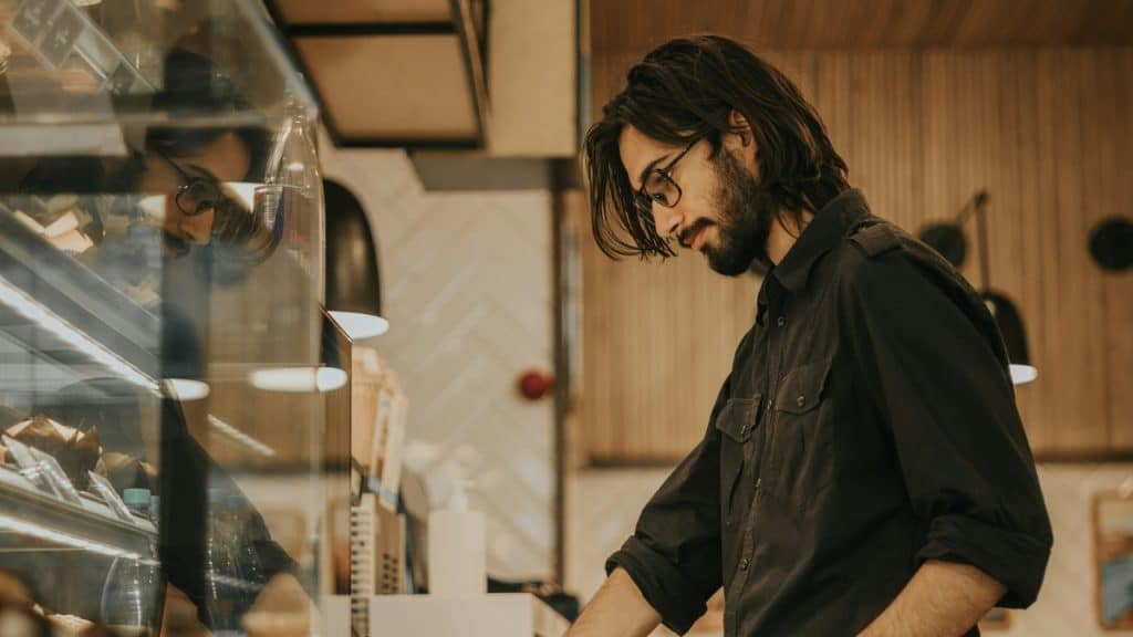 A man with long hair and glasses looks into a glass display case in a shop.