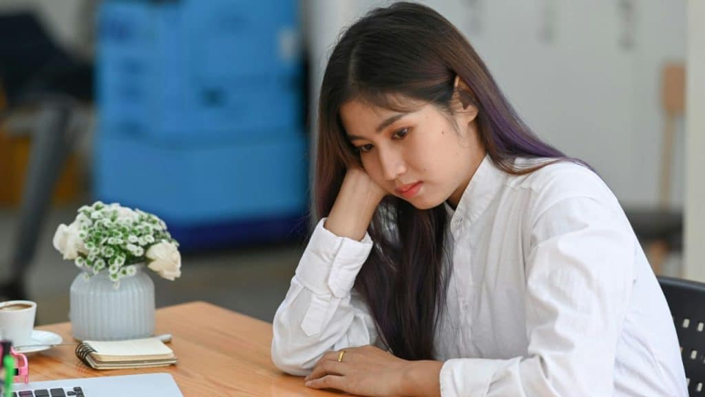 A woman in a white shirt with purple highlights in her hair rests her head on her hand at a desk.