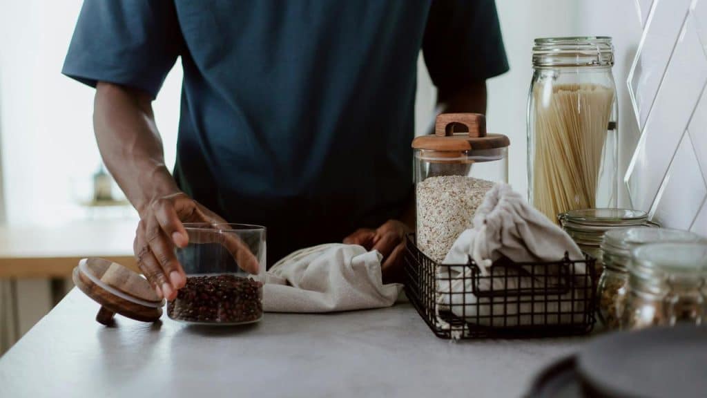A person placing beans into a glass container on a kitchen counter.