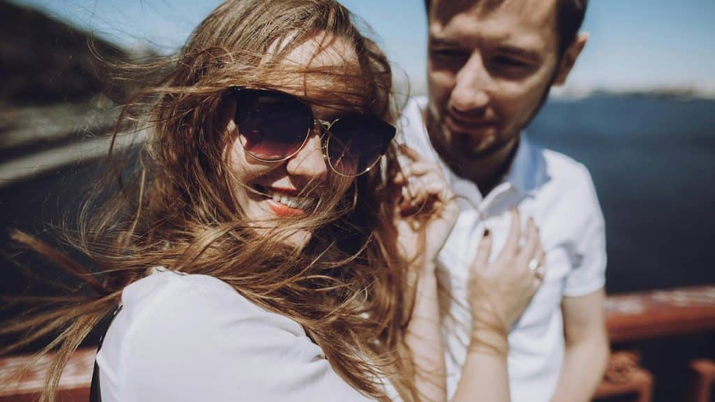 A smiling woman with sunglasses and a man standing together on a windy day by the water.