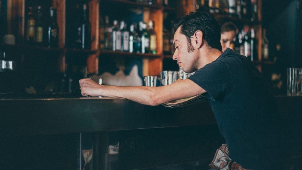 A man in a dark t-shirt leans over a bar counter in a dimly lit room.