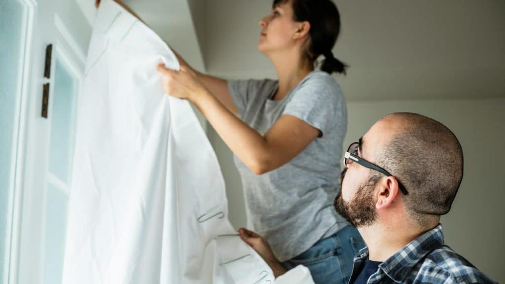 A person hanging a white curtain while another person looks on nearby.