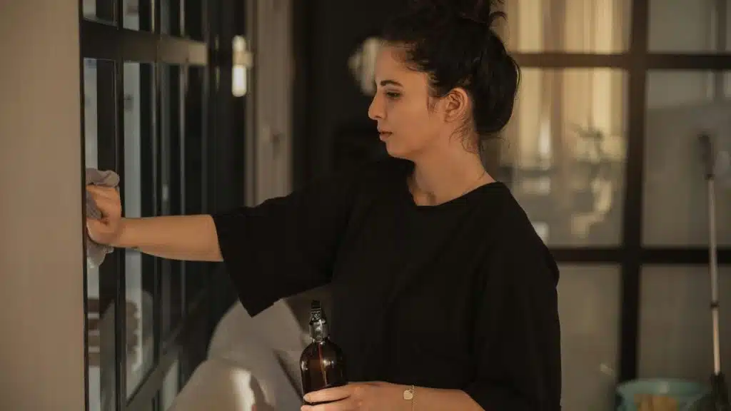 A woman wipes a glass surface while holding a spray bottle indoors.