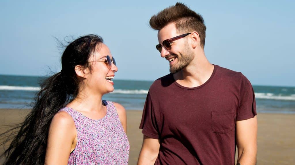 A couple wearing sunglasses smiling at each other while walking on the beach.