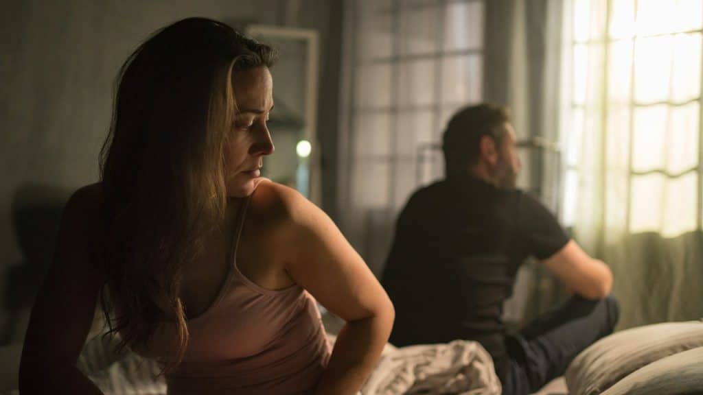 A woman and man sitting with their backs to each other and not talking while they sit on a bed.