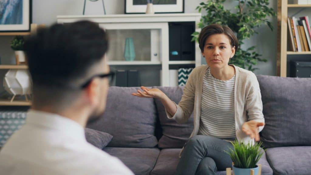 A woman sits on a couch gesturing with her hands while speaking to a man.