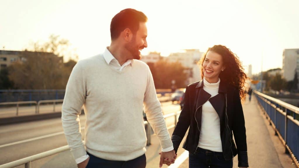 A man and woman holding hands and smiling while walking outdoors during a bright sunset.