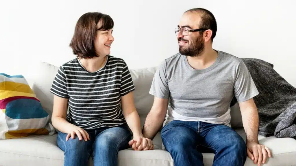 A man and woman sit on a sofa, holding hands and smiling at each other.