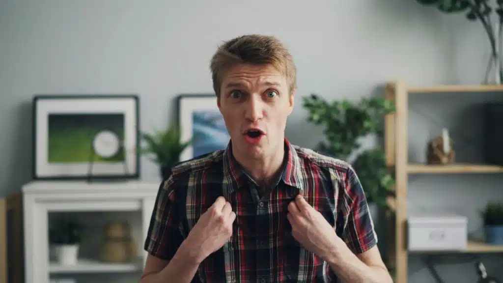 A man in a plaid shirt gestures toward himself with a wide-eyed, surprised expression.