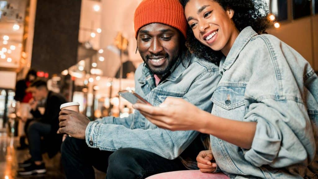 A smiling couple sitting together indoors, looking at a phone while holding coffee.