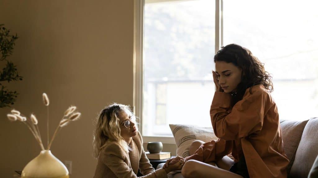 A person sitting on a couch with head bowed while another person offers comfort nearby.