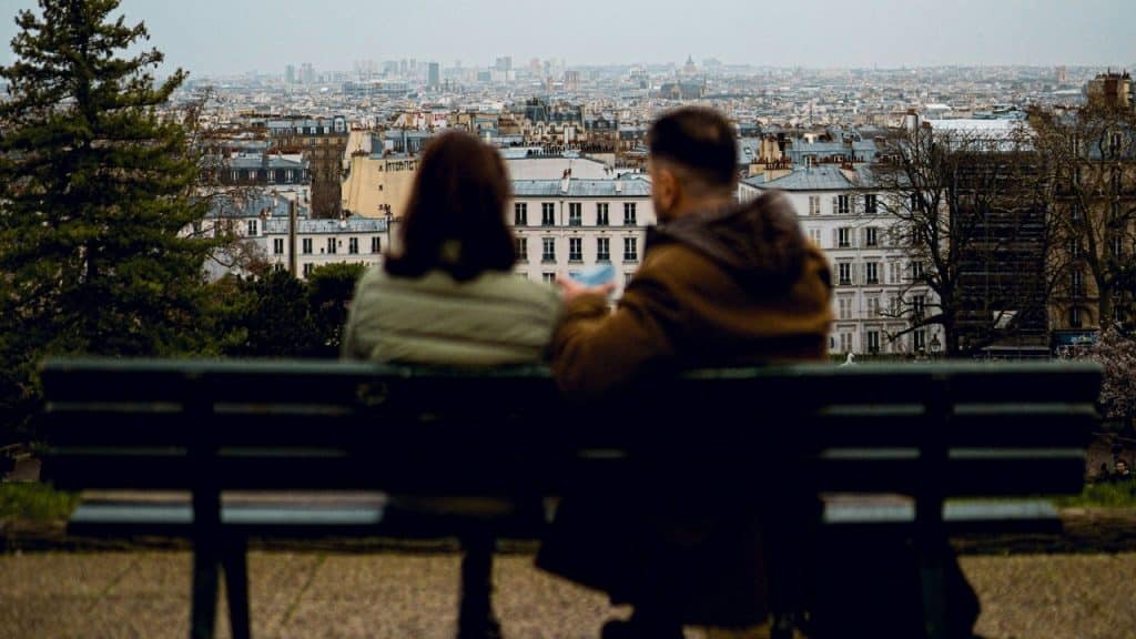 A man and woman sit on a park bench looking out over a city skyline.