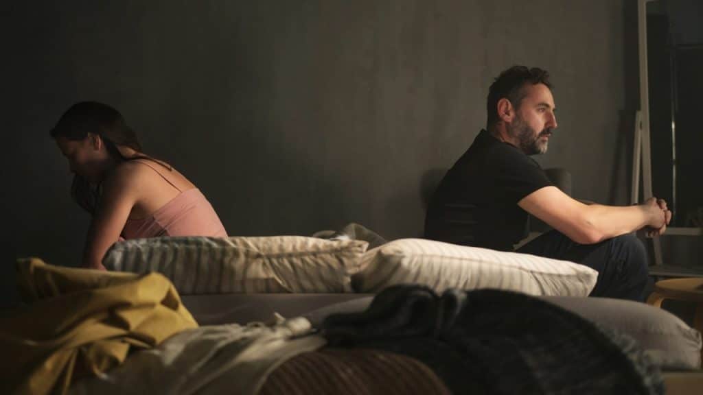 A man and woman sit on the edge of a bed facing away from each other.