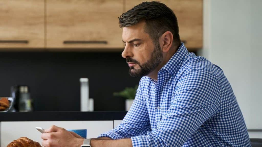 A man in a blue checkered shirt sits at a table and looks at a smartphone.
