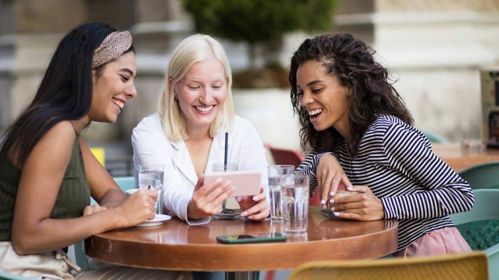 A group of friends laughing together while looking at a phone at an outdoor café.