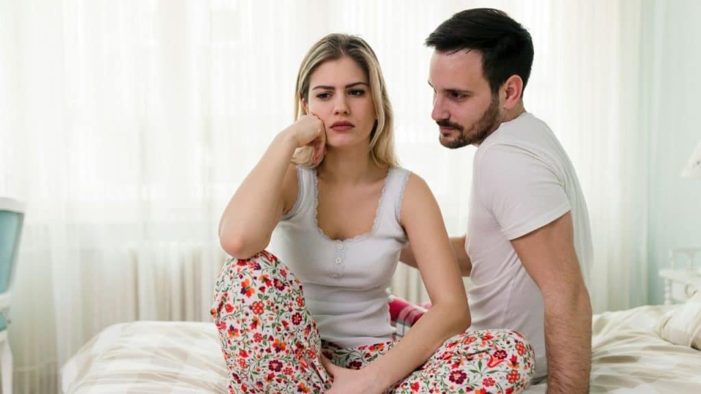 A man in a white shirt looks at a woman sitting cross-legged on a bed.