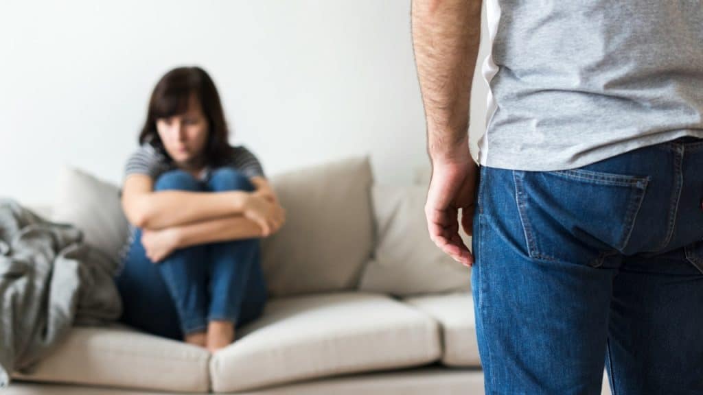 A woman sits on a sofa with her knees tucked while a man stands in foreground.