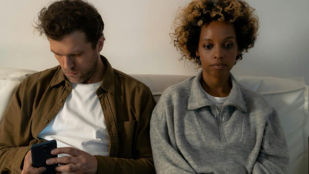 A man and woman sit next to each other on a white sofa, both looking downward.